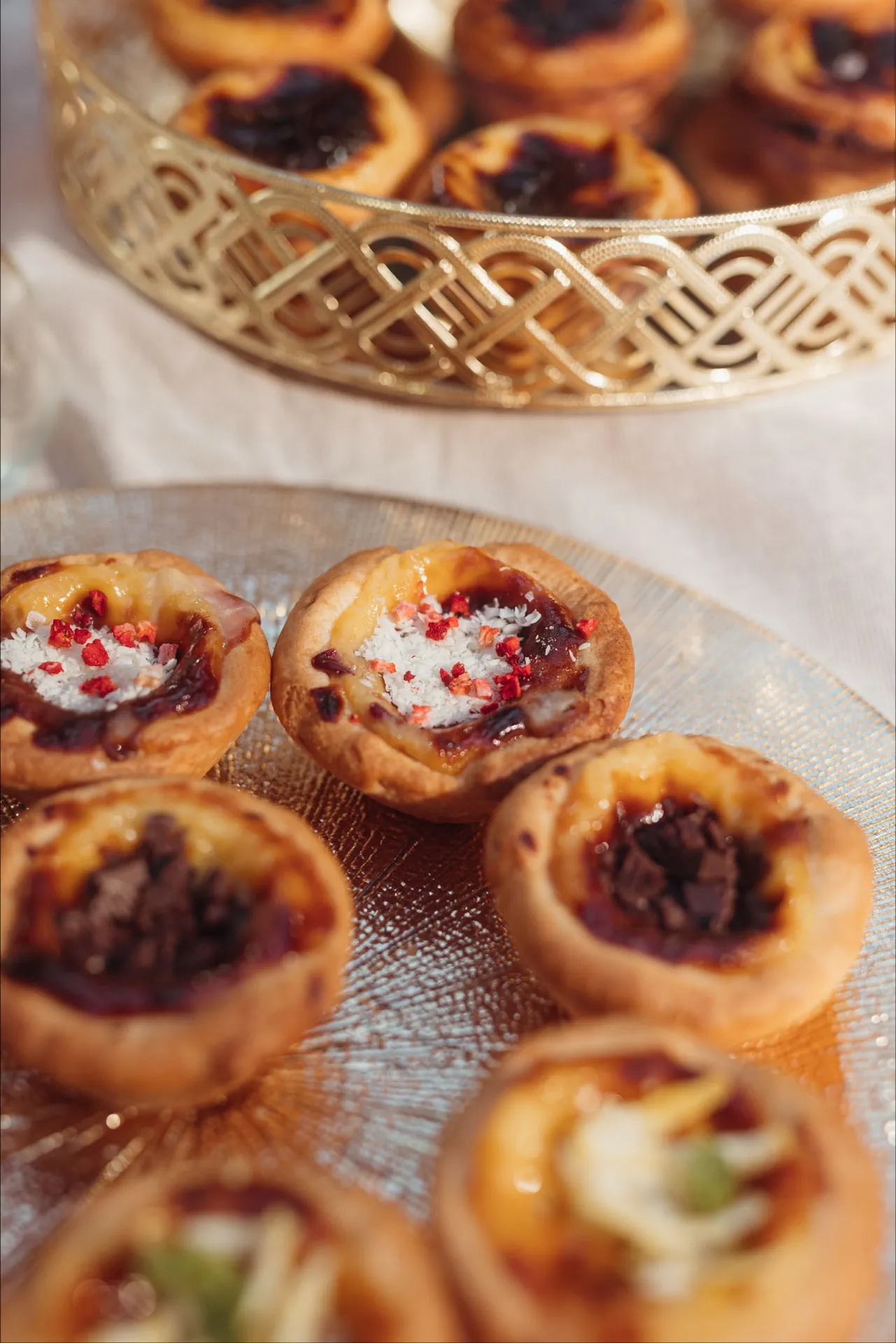 Babeczki z maliną, białą posypką i czekoladą na szklanym talerzu, za nimi Pastéis de Nata w złotym koszyku