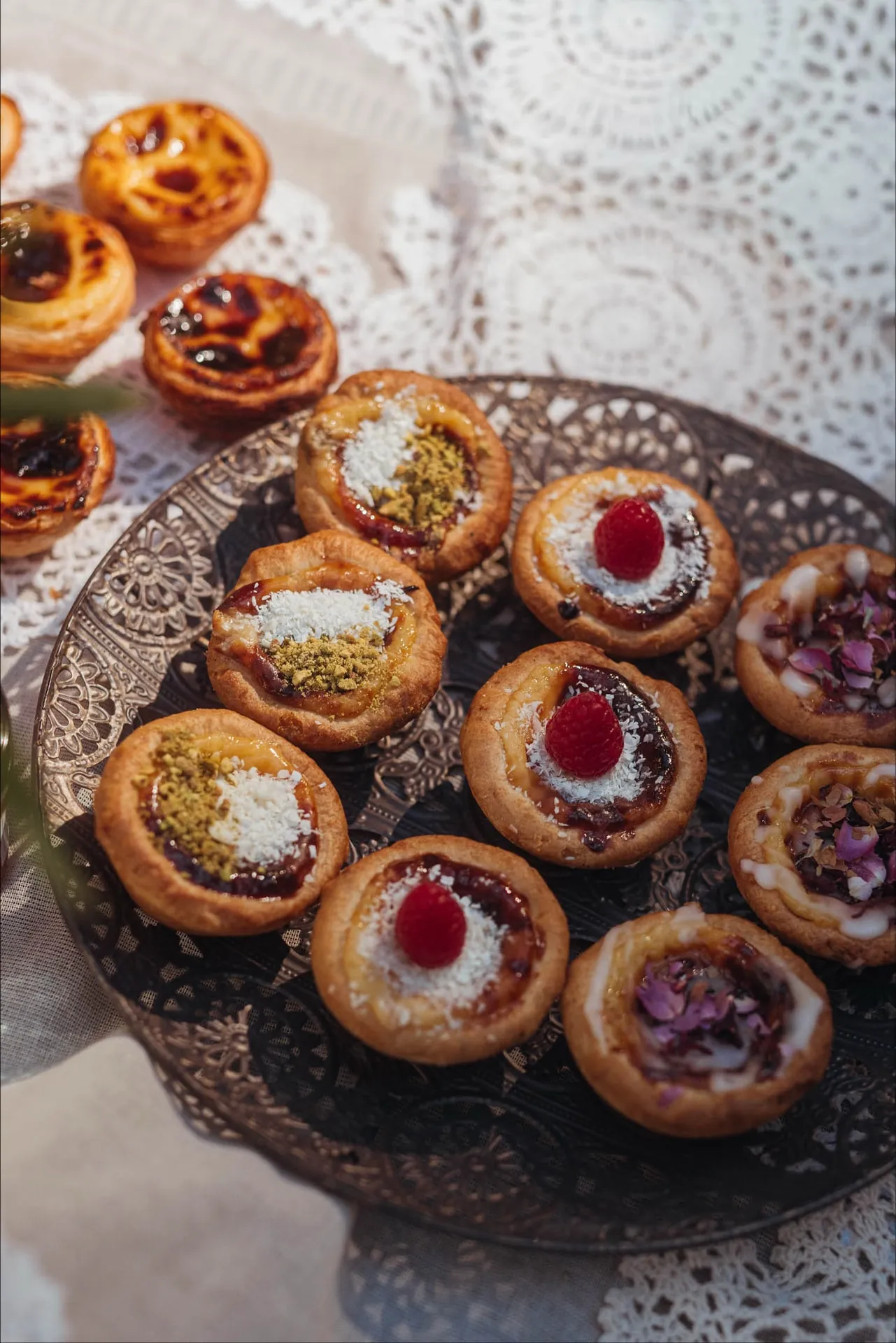 Babeczki BOM DIA z maliną, pistacjami i płatkami kwiatów na ażurowej tacy, w tle Pastéis de Nata na koronkowym obrusie