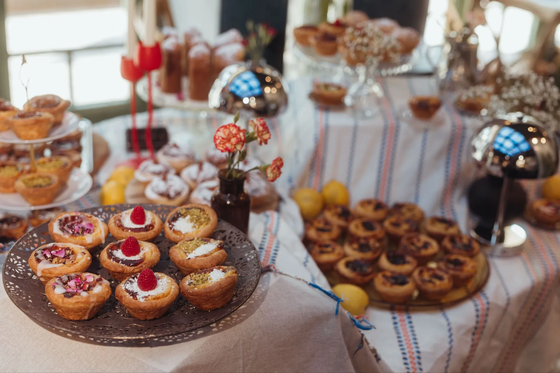 Słodki stół na pasiastym obrusie: Pastéis de Nata i babeczki BOM DIA na tacach, cytryny i kwiaty, w tle inne desery