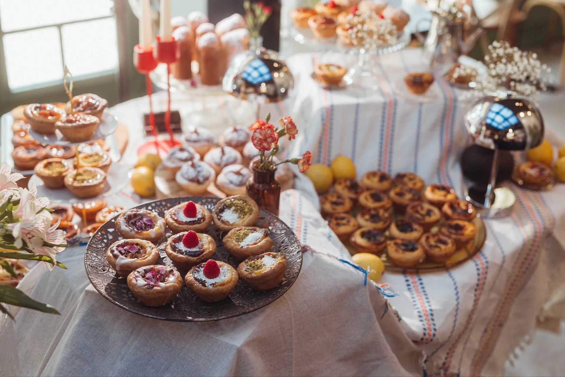 Słodki stół z Pastéis de Nata, babeczkami BOM DIA, babkami i cytrynami na pasiastym obrusie, obok kwiaty