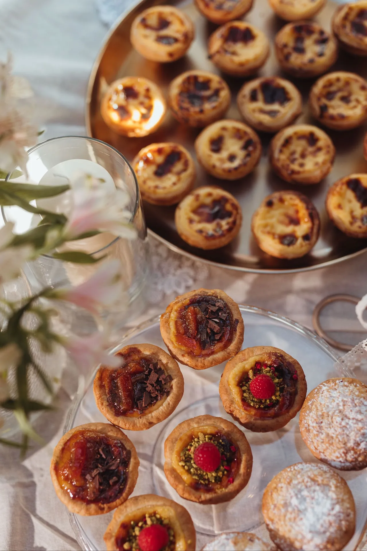 Okrągła taca z Pastéis de Nata, przed nią szklany talerz z babeczkami BOM DIA i ciastkami z cukrem pudrem