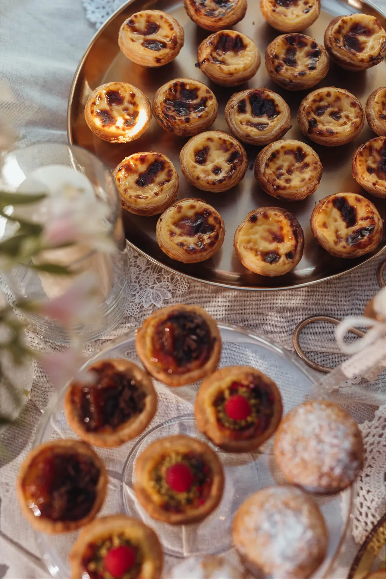 Okrągła taca pełna Pastéis de Nata, przed nią babeczki BOM DIA i ciastka z cukrem pudrem na szklanym talerzu