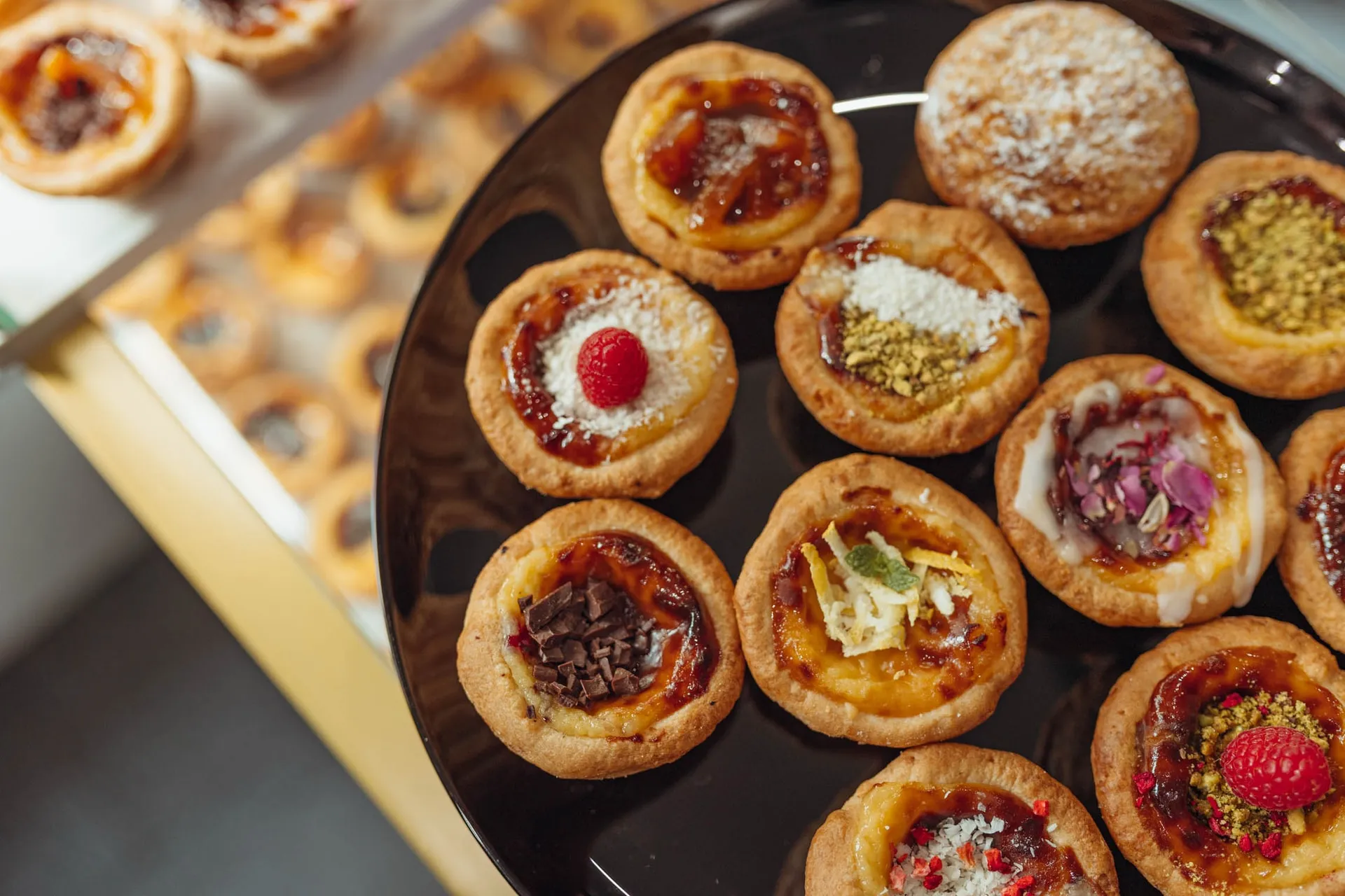Czarna taca autorskich babeczek BOM DIA z maliną, pistacjami, czekoladą i cukrem pudrem, w tle Pastéis de Nata w witrynie
