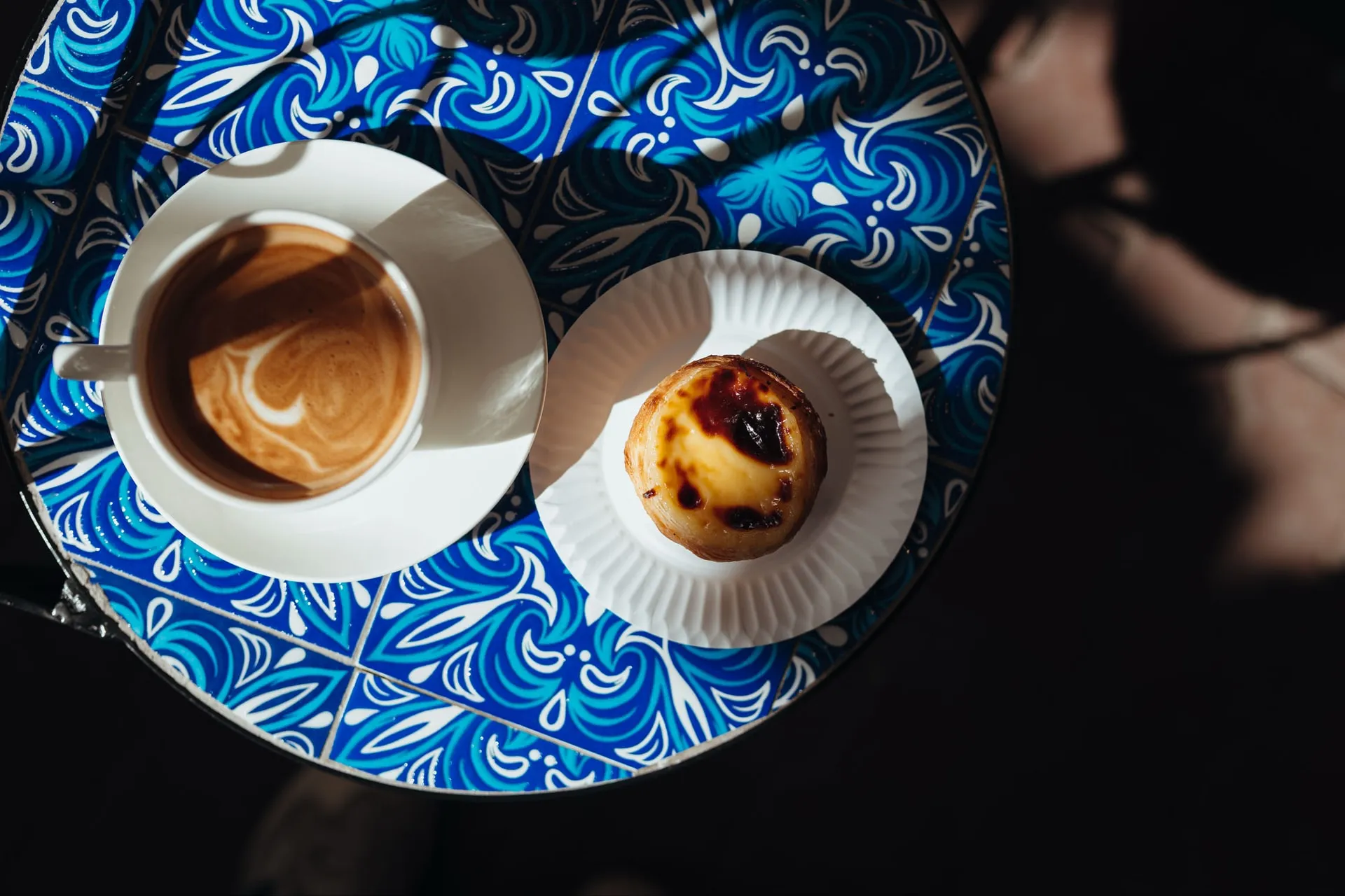 Widok z góry: filiżanka kawy z mlecznym wzorem i Pastel de Nata na papierowej podkładce, na stoliku w niebieskie wzory