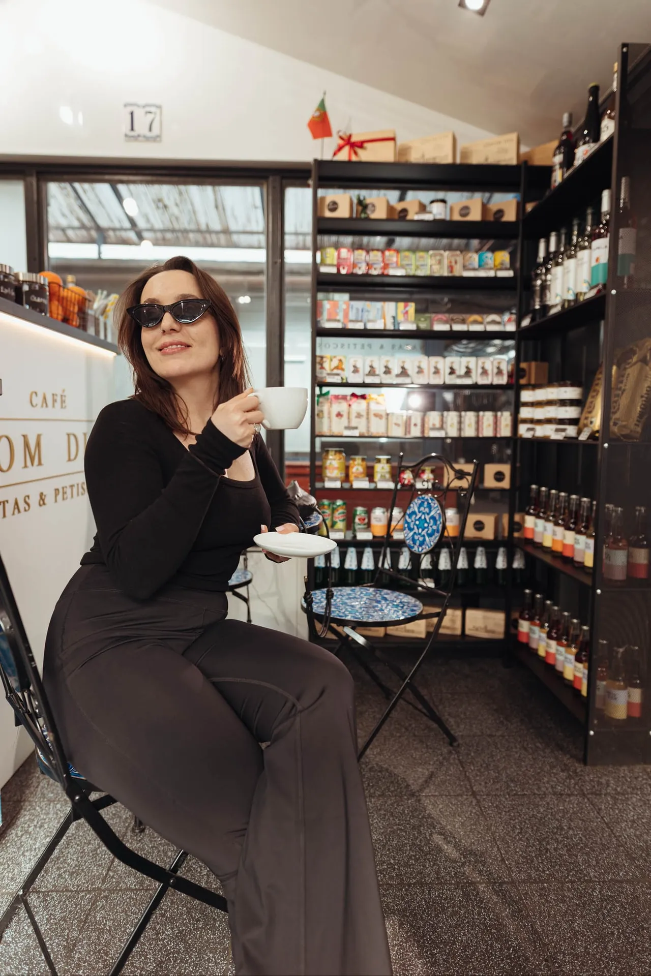 Kobieta w okularach przeciwsłonecznych pije kawę przy kiosku 17, za nią półki z delikatesami i winem