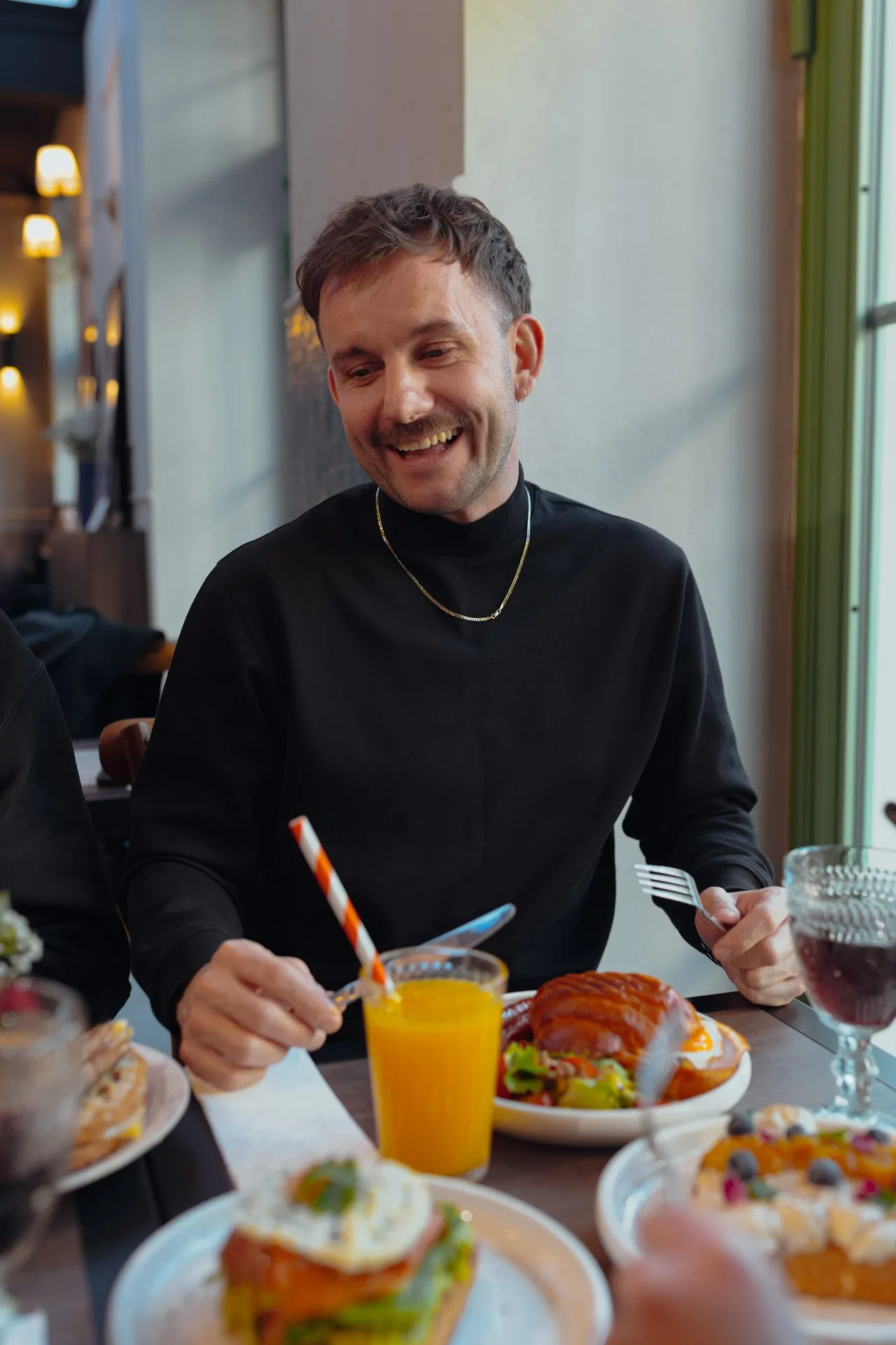 Roześmiany mężczyzna w czarnym golfie przy stole ze śniadaniem: rogal, sok pomarańczowy z pasiastą słomką, kieliszek z czerwonym napojem i tost z jajkiem sadzonym