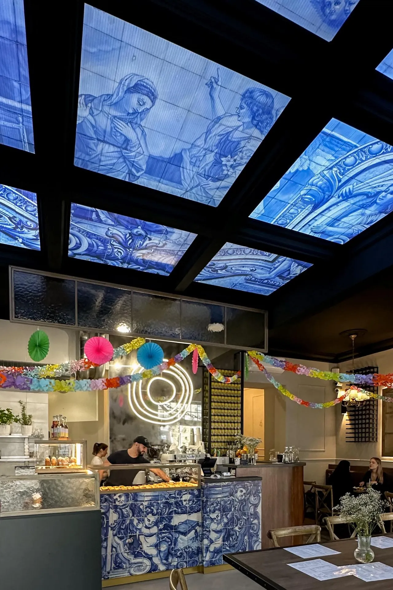 Podświetlony sufit z niebieskich paneli azulejos z postaciami i ornamentami nad ladą BOM DIA przy Krakowskiej; pod nim kolorowe girlandy, neon z logo i lada wyłożona azulejos