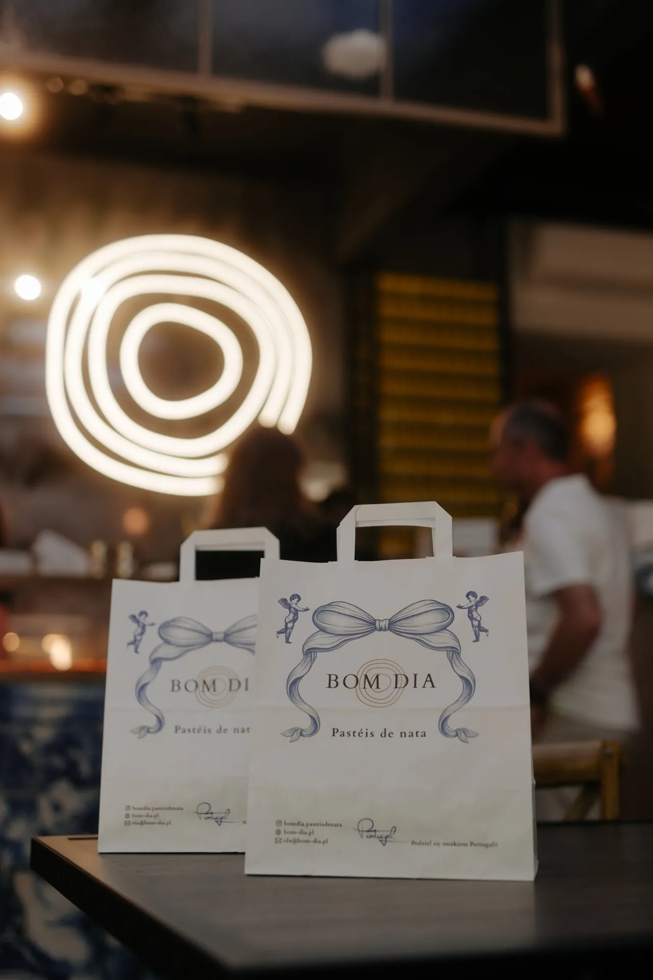Dwie papierowe torby BOM DIA Pastéis de Nata na stole, w tle neon z logo i goście w lokalu