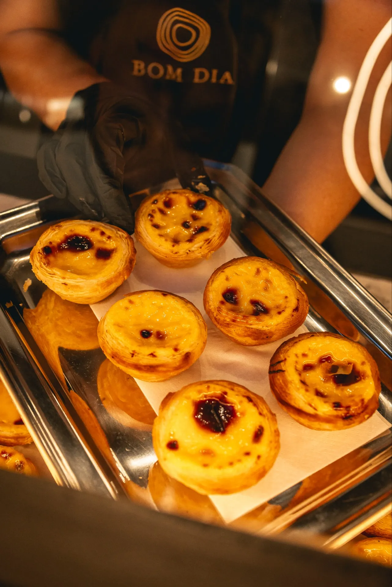 Pastéis de Nata na metalowej blasze, którą cukiernik w czarnej rękawiczce wysuwa w stronę gości; w tle fartuch z logo BOM DIA