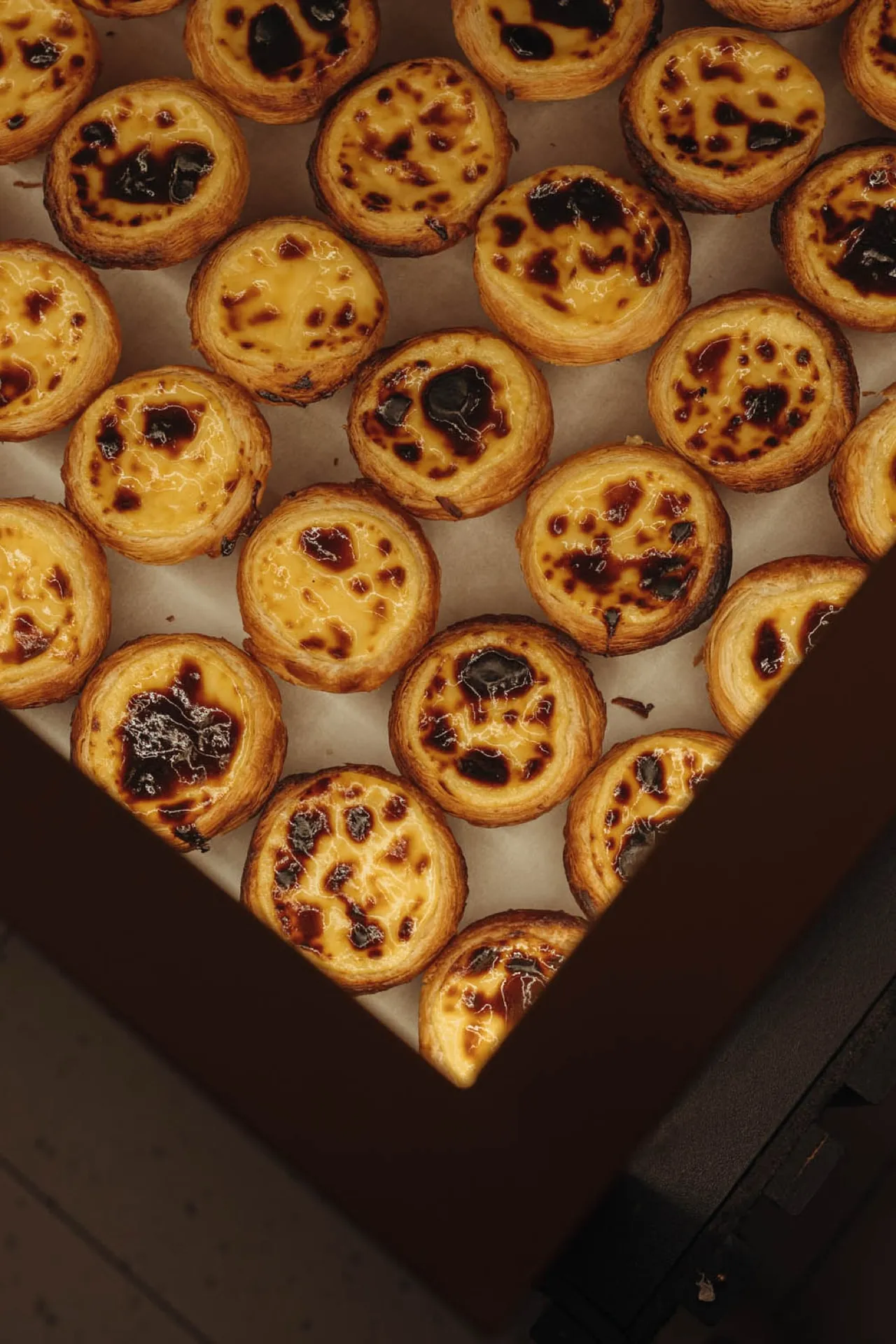 Pastéis de Nata z przypieczonymi plamkami na białym papierze, widziane z góry, w narożniku ciemnobrązowego pojemnika