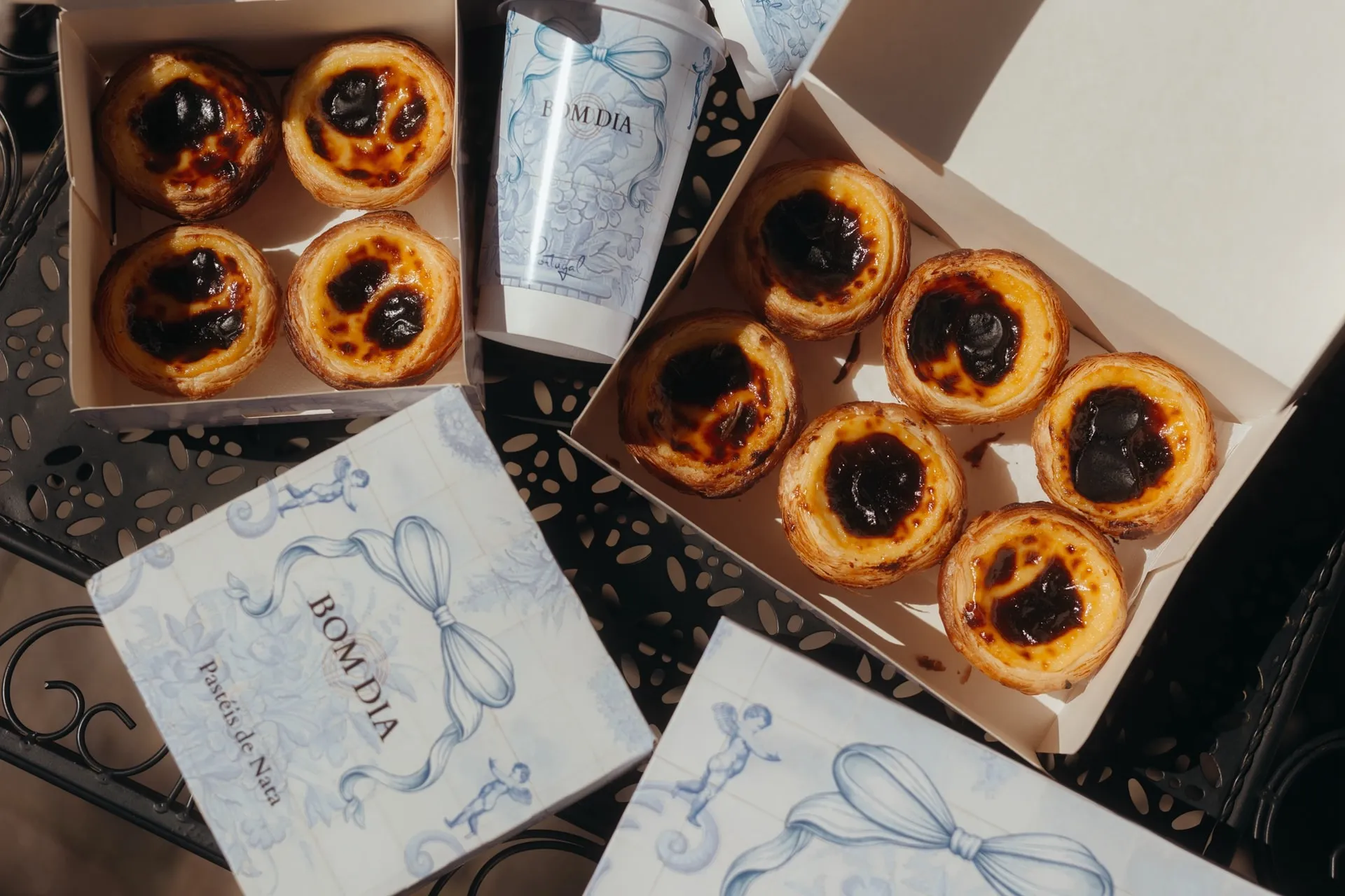 Pastéis de Nata w dwóch otwartych pudełkach BOM DIA i kawa na wynos, obok zamknięte pudełka z niebieskim wzorem