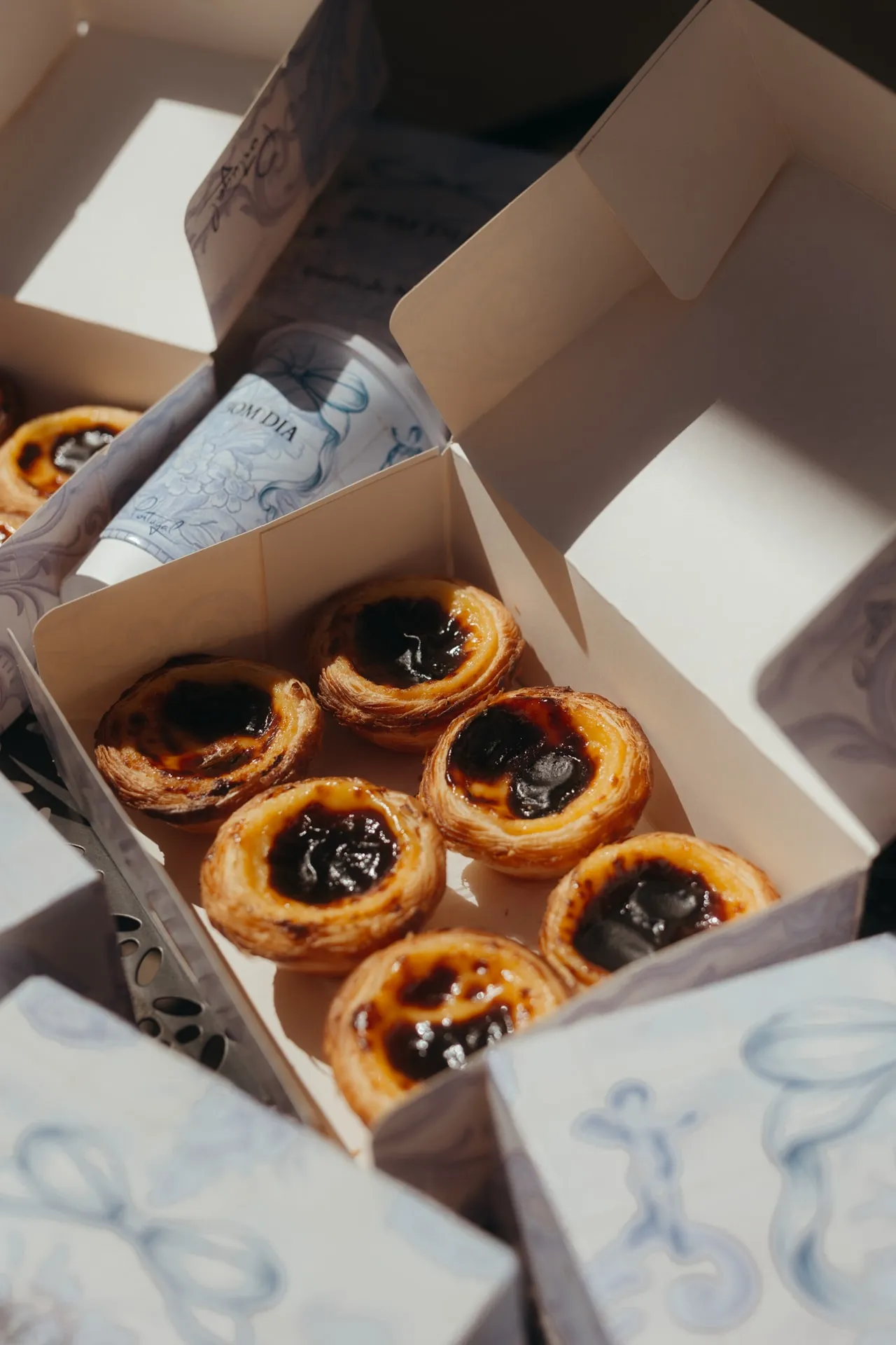 Otwarte pudełko z sześcioma Pastéis de Nata, obok kubek BOM DIA i pudełka z niebieskim wzorem kokardy
